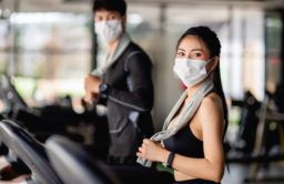 Selective focus,  young sexy woman in mask wearing sportwear and smartwatch and blurred young man on background, They are running on treadmill to for workout in modern gym, look at camera, copy space